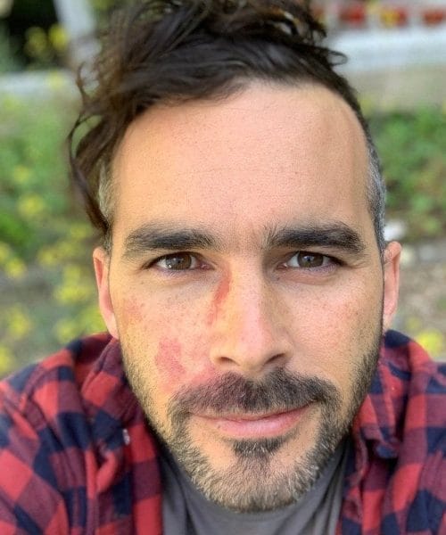 A man with dark hair and a beard, wearing a red and black plaid shirt, looks directly at the camera with a slight smile. The background is outdoors with blurred greenery and yellow flowers.