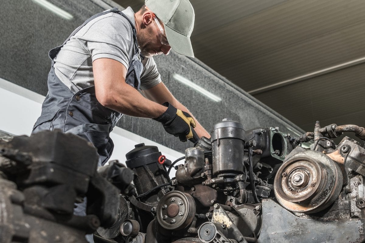 A mechanic wearing a cap, gloves, and work clothes repairs a car engine in a garage, using a wrench to adjust parts under bright indoor lighting.