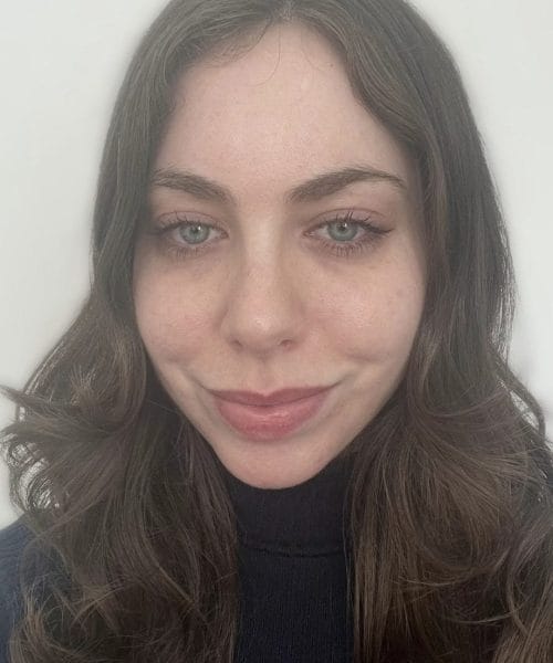 A woman with wavy brown hair and light eyes, wearing a dark turtleneck sweater, smiles softly against a plain white background.