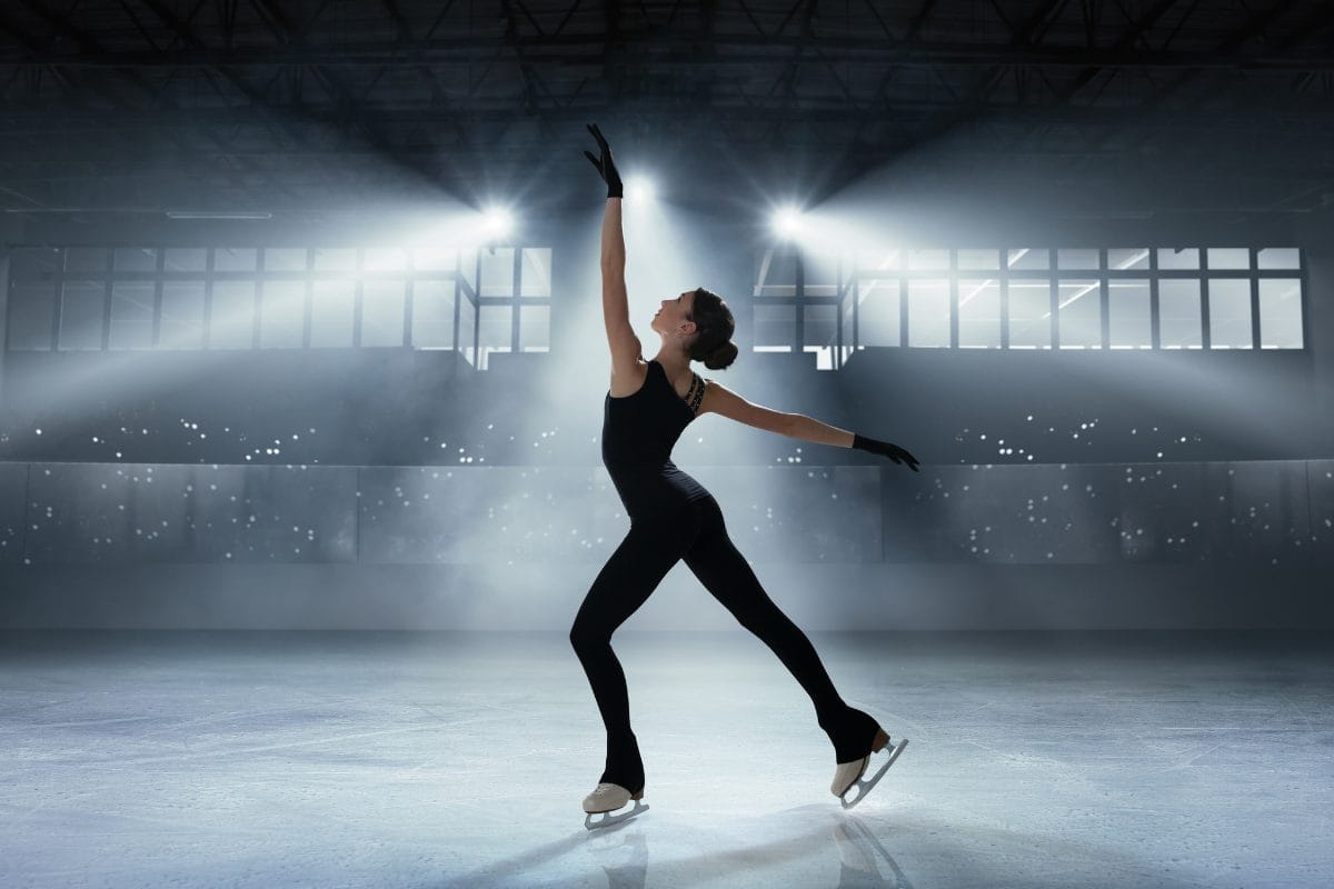 A figure skater in a black outfit poses gracefully on an ice rink, one arm raised and one leg extended behind, with bright spotlights and a large windowed wall in the background.