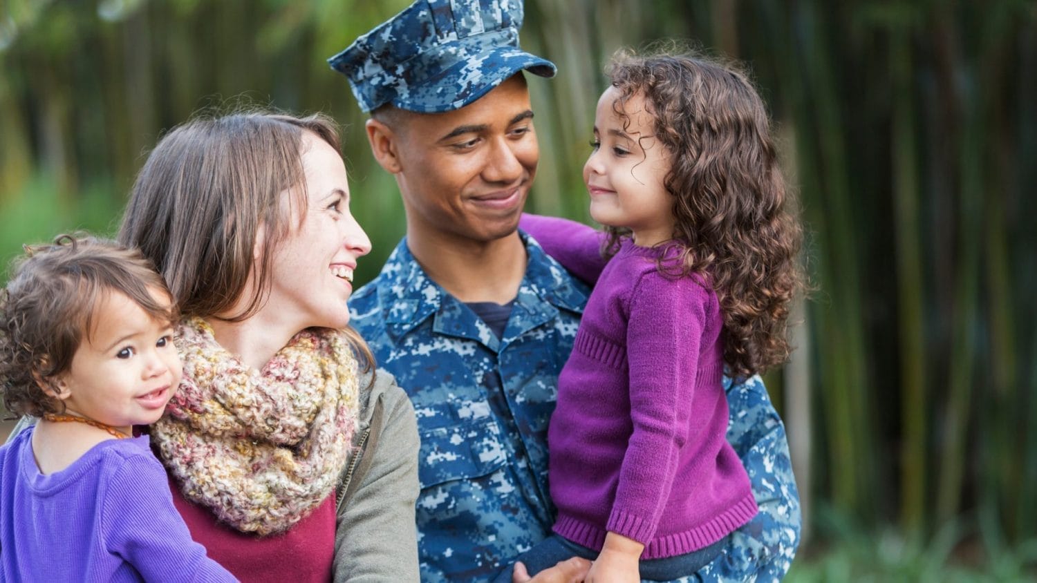 A smiling family stands outdoors. A man in a navy blue military uniform holds a young girl in a purple sweater, while a woman with a scarf holds another young girl, all looking happy and close together.